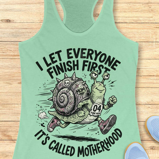 Light green tank top with cartoon snail and text I let everyone finish first, It's called motherhood, on a wooden surface