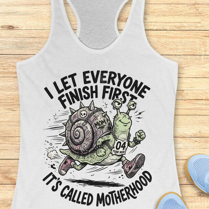 white tank top with humorous graphic and text  I let everyone finish first, It's called motherhood, on a wooden surface