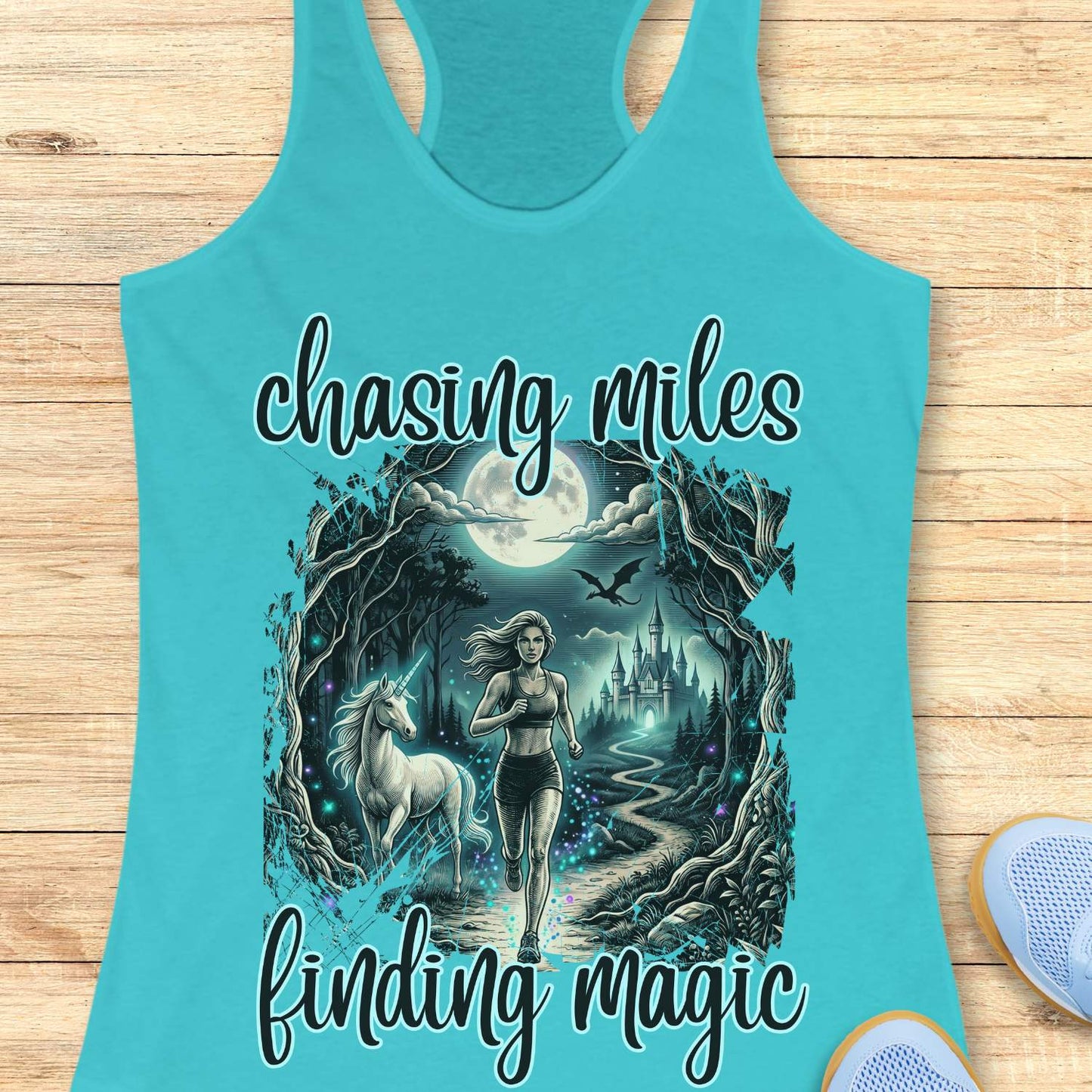 Turquoise tank top with a graphic design of a woman and unicorn, text 'chasing miles finding magic', on a wooden surface.