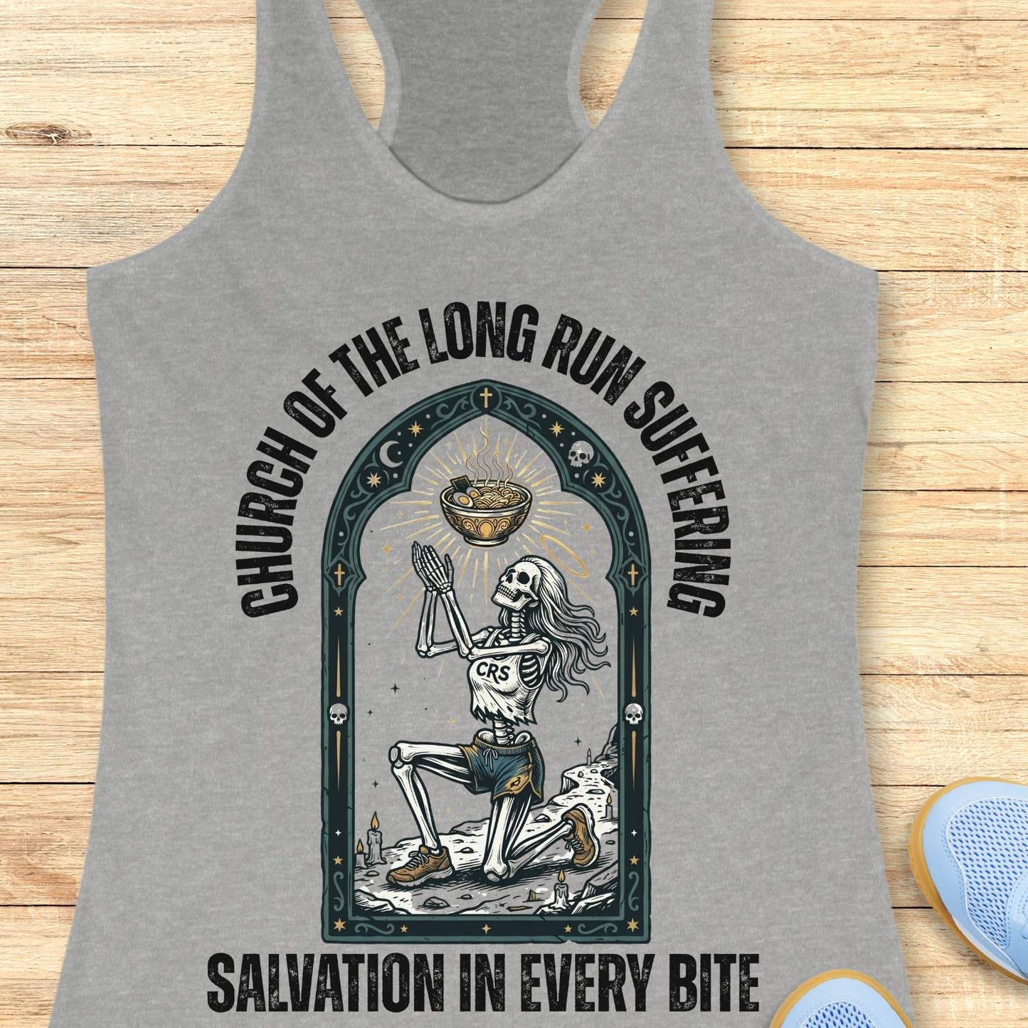 Gray tank top with skeleton design and text 'Church of the long run suffering. Salvation in every bite', on a wooden surface