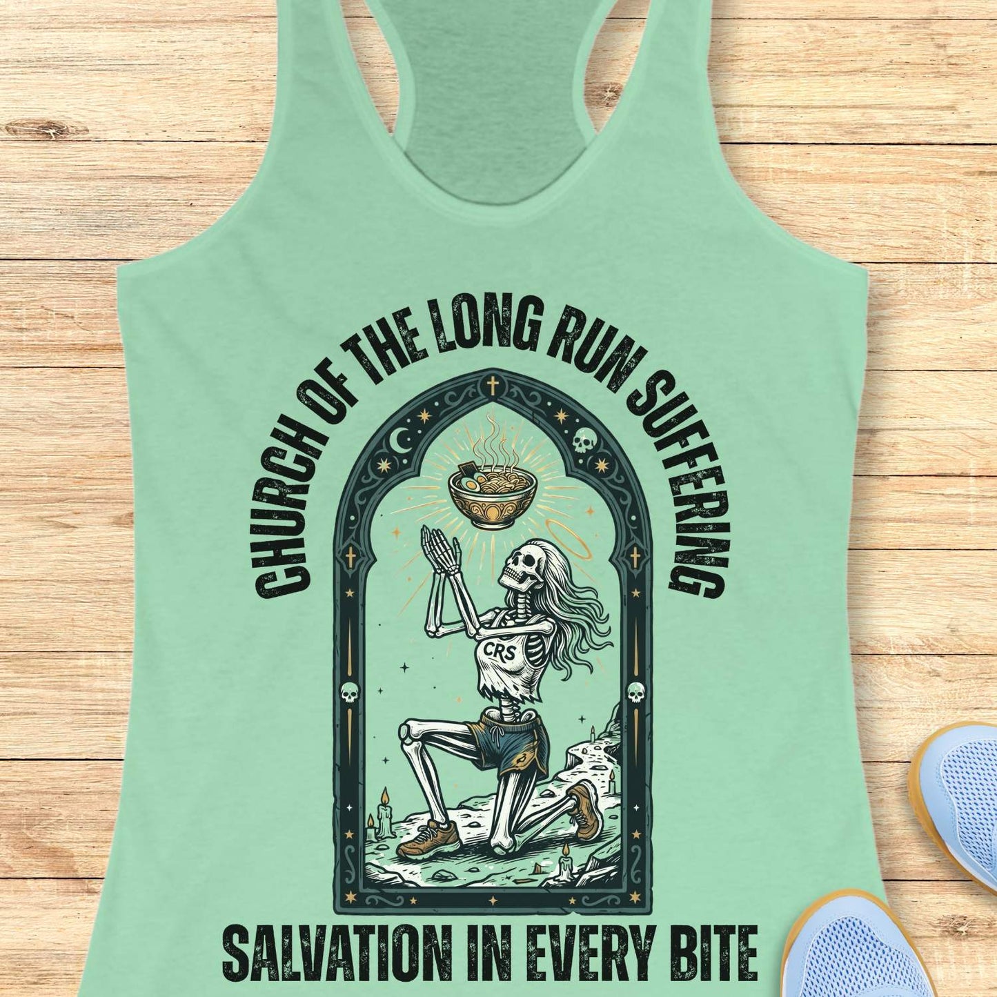 Mint green tank top with skeleton design and text 'Church of the long run suffering. Salvation in every bite', on a wooden surface