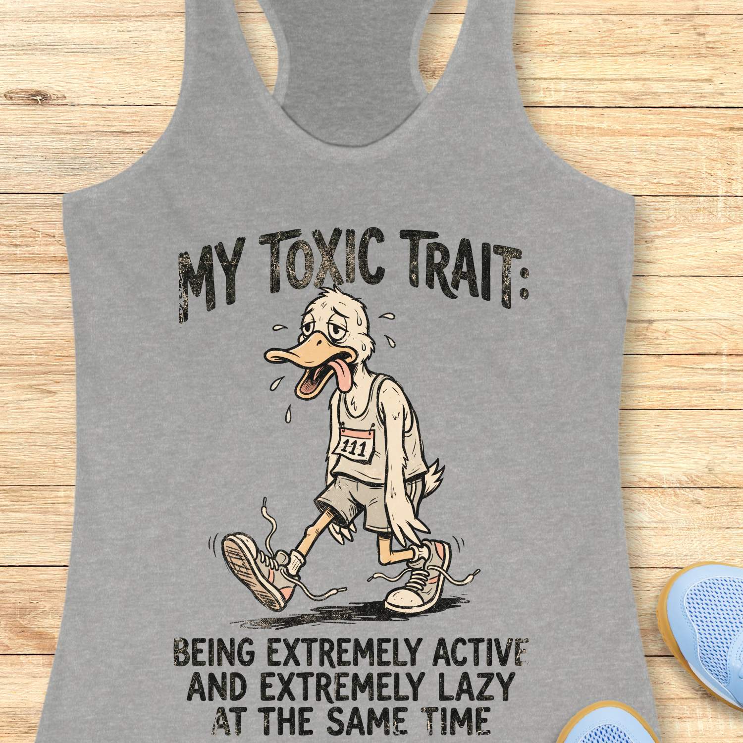 Gray tank top with cartoon character and humorous text ' my toxic trait - being extremely active and extremely lazy at the same time',  on a wooden surface