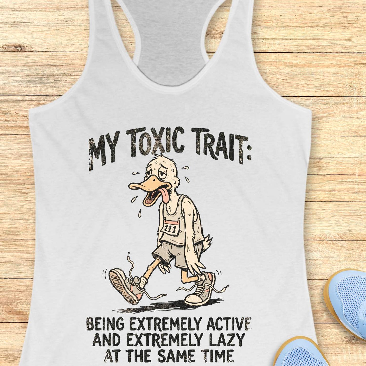 white tank top with cartoon character and text ' my toxic trait - being extremely active and extremely lazy at the same time',  on a wooden surface