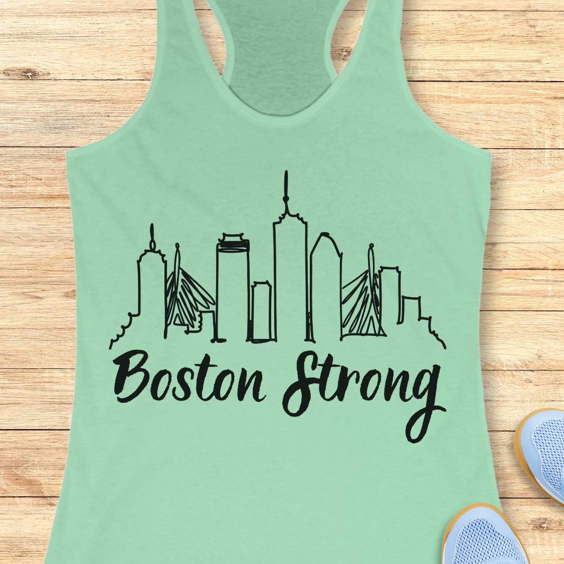 Light green tank top with 'Boston Strong' and city skyline design on a wooden surface.