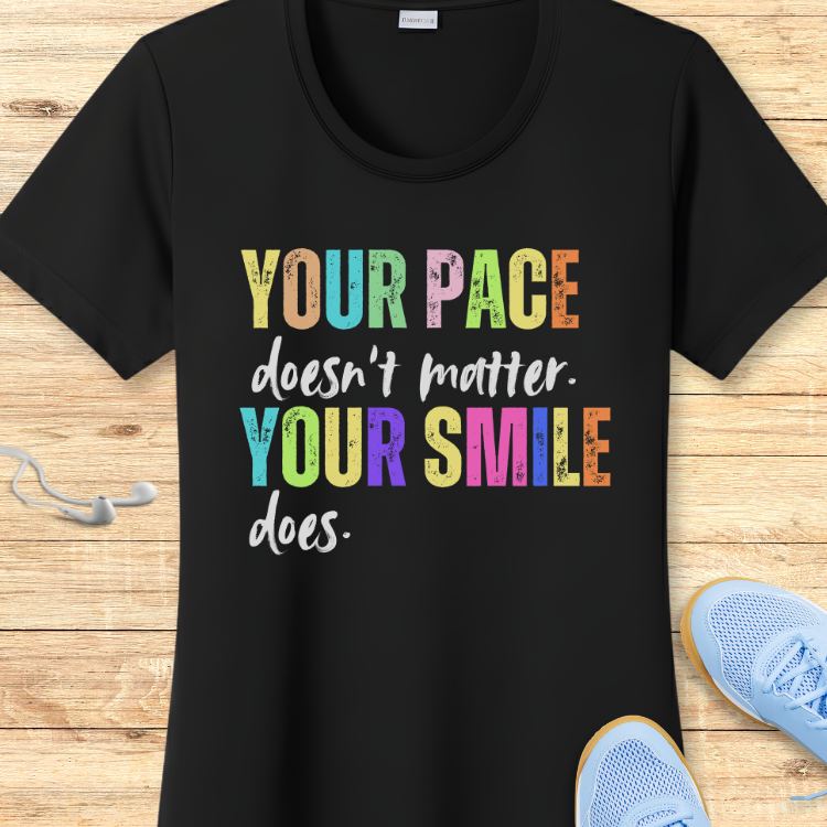 Black t-shirt displays the quote YOUR PACE doesn't matter. YOUR SMILE does with YOUR PACE and YOUR SMILE in vibrant multicolor block letters, surrounded by white wired earbuds and light blue mesh athletic shoes on a wooden background