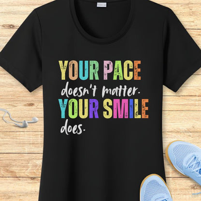 Black t-shirt displays the quote YOUR PACE doesn't matter. YOUR SMILE does with YOUR PACE and YOUR SMILE in vibrant multicolor block letters, surrounded by white wired earbuds and light blue mesh athletic shoes on a wooden background