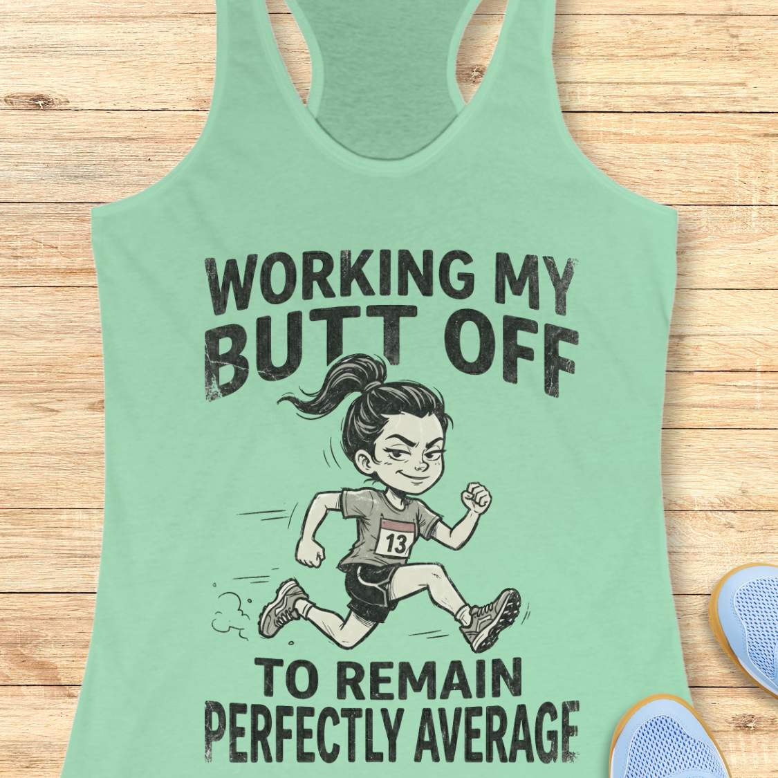 Mint green tank top with a graphic and text  'working my butt off to remain perfectly average', on a wooden surface