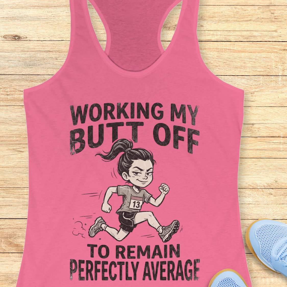 Pink tank top with a graphic and text  'working my butt off to remain perfectly average', on a wooden surface