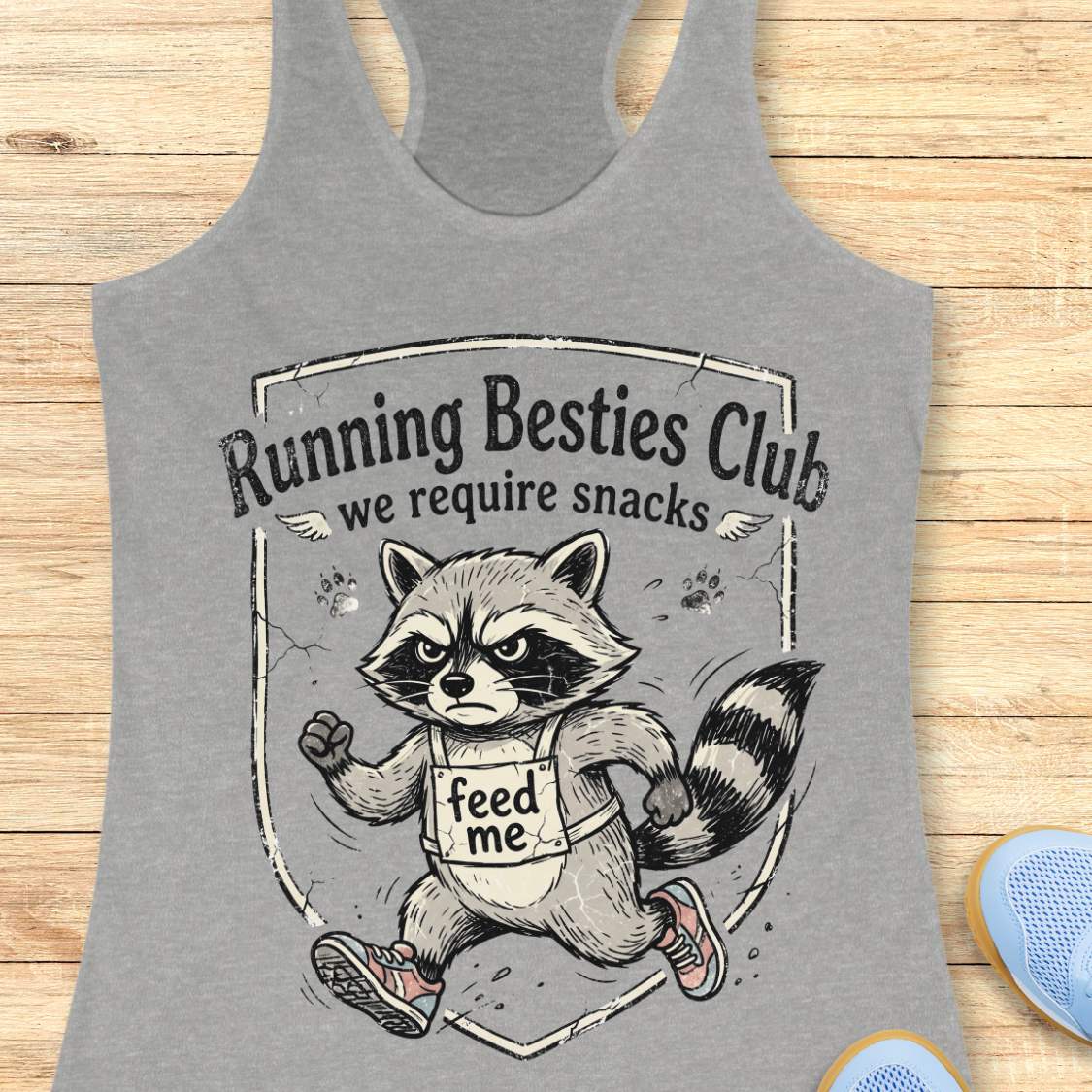 Gray tank top with raccoon graphic and 'Running Besties Club, we require snacks' text on a wooden surface