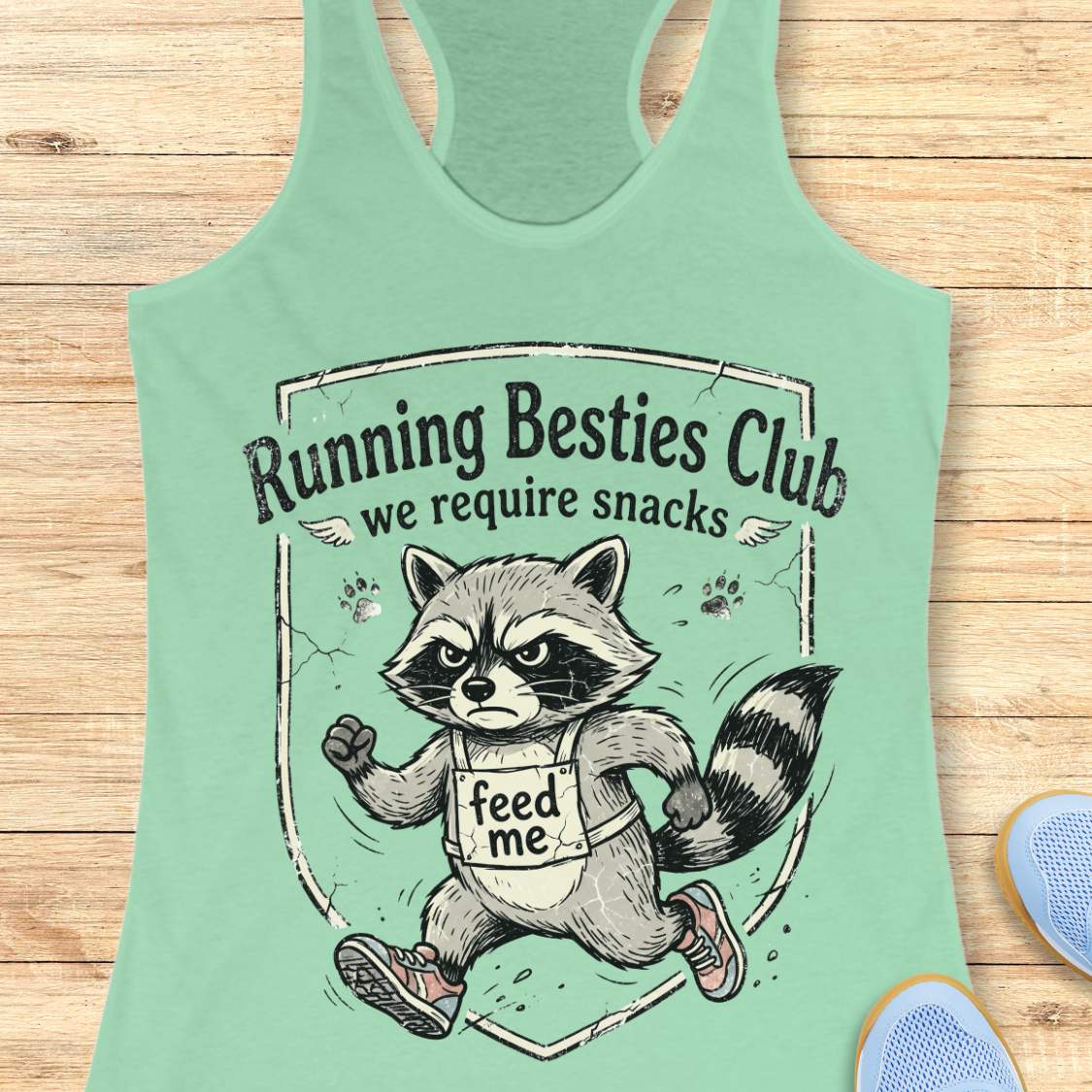Mint green tank top with raccoon graphic and text 'Running Besties Club, we require snacks' on a wooden surface.