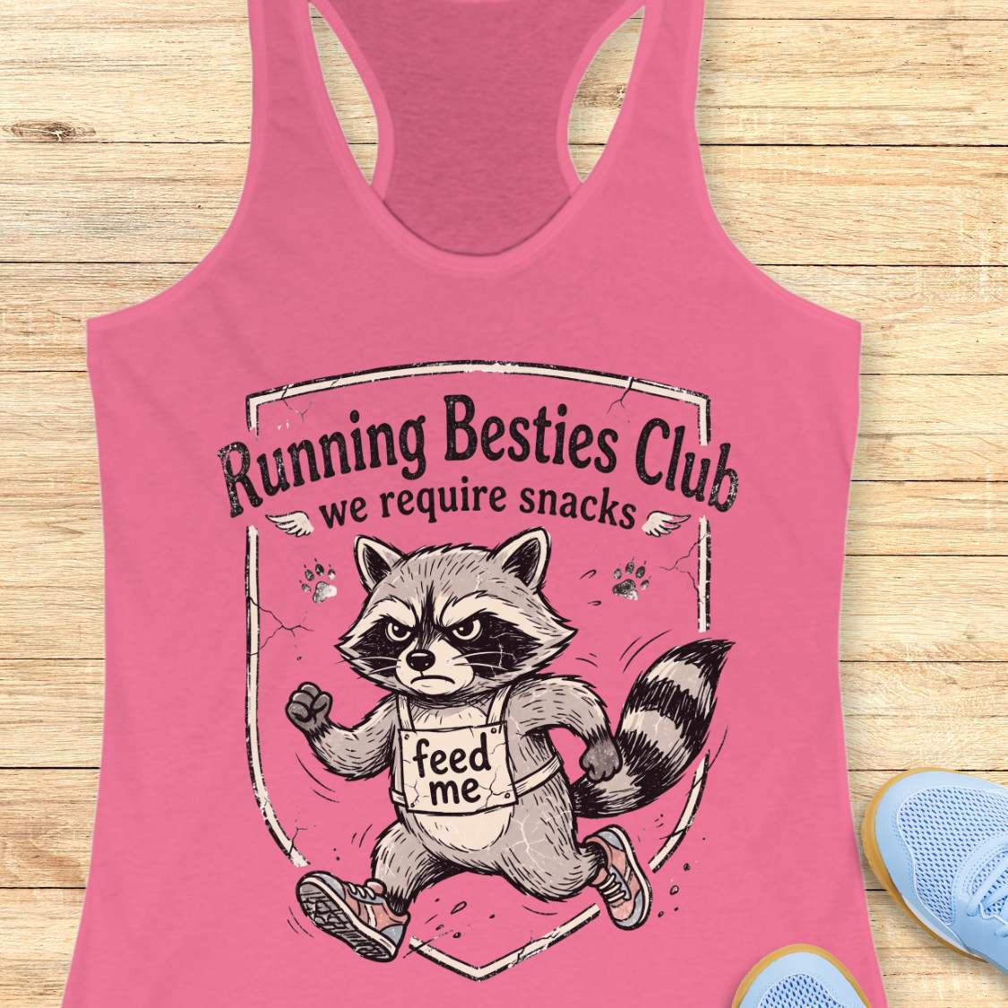 Pink tank top with raccoon graphic and text 'Running Besties Club, we require snacks' on a wooden surface.