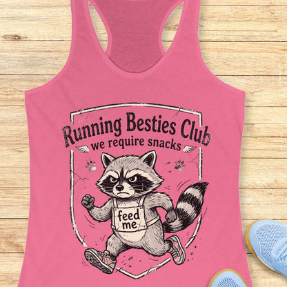 Pink tank top with raccoon graphic and text 'Running Besties Club, we require snacks' on a wooden surface.