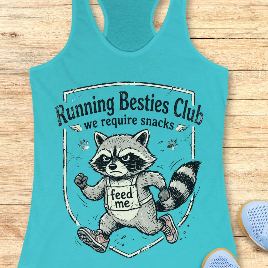 Turquoise tank top with raccoon graphic and text 'Running Besties Club, we require snacks' on a wooden surface.