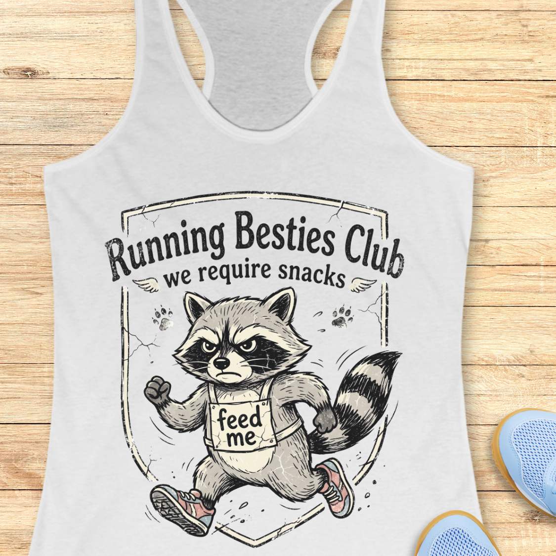white tank top with a raccoon graphic and text 'Running Besties Club, we require snacks' on a wooden surface.