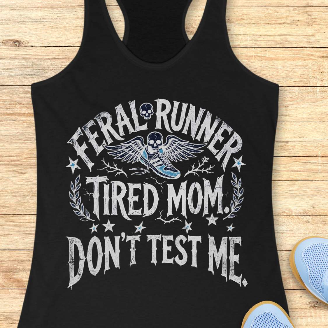 Black tank top with white text 'feral runner, tired mom. Don't test me.',  and graphic on a wooden surface
