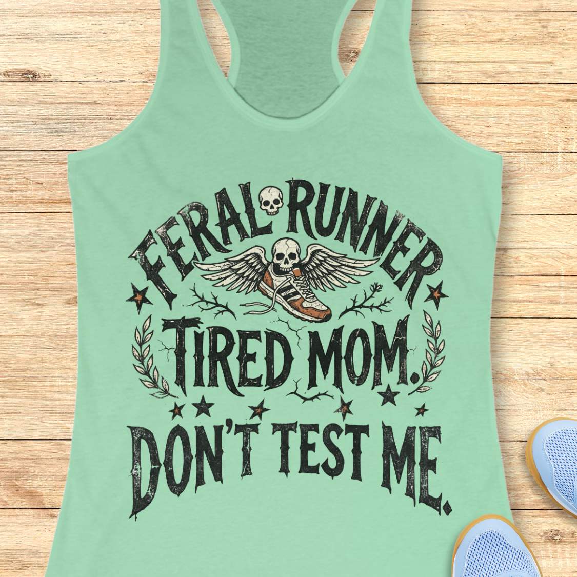 Mint green tank top with text 'feral runner, tired mom. Don't test me.', and graphic design on a wooden surface