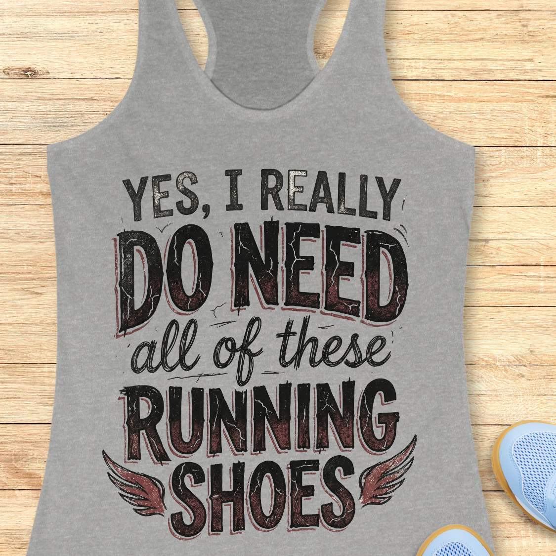 Gray tank top with motivational text ' yes, I really do need all of these running shoes',  on a wooden surface