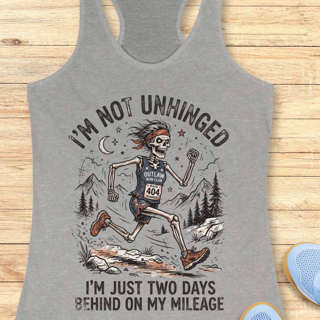 Gray tank top with skeleton runner graphic and text 'I'm not unhinged, I'm just two days behind on my mileage', on a wooden surface