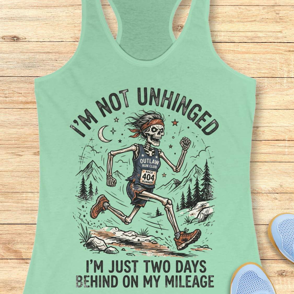 Mint green tank top with skeleton graphic and text 'I'm not unhinged, I'm just two days behind on my mileage', on a wooden surface