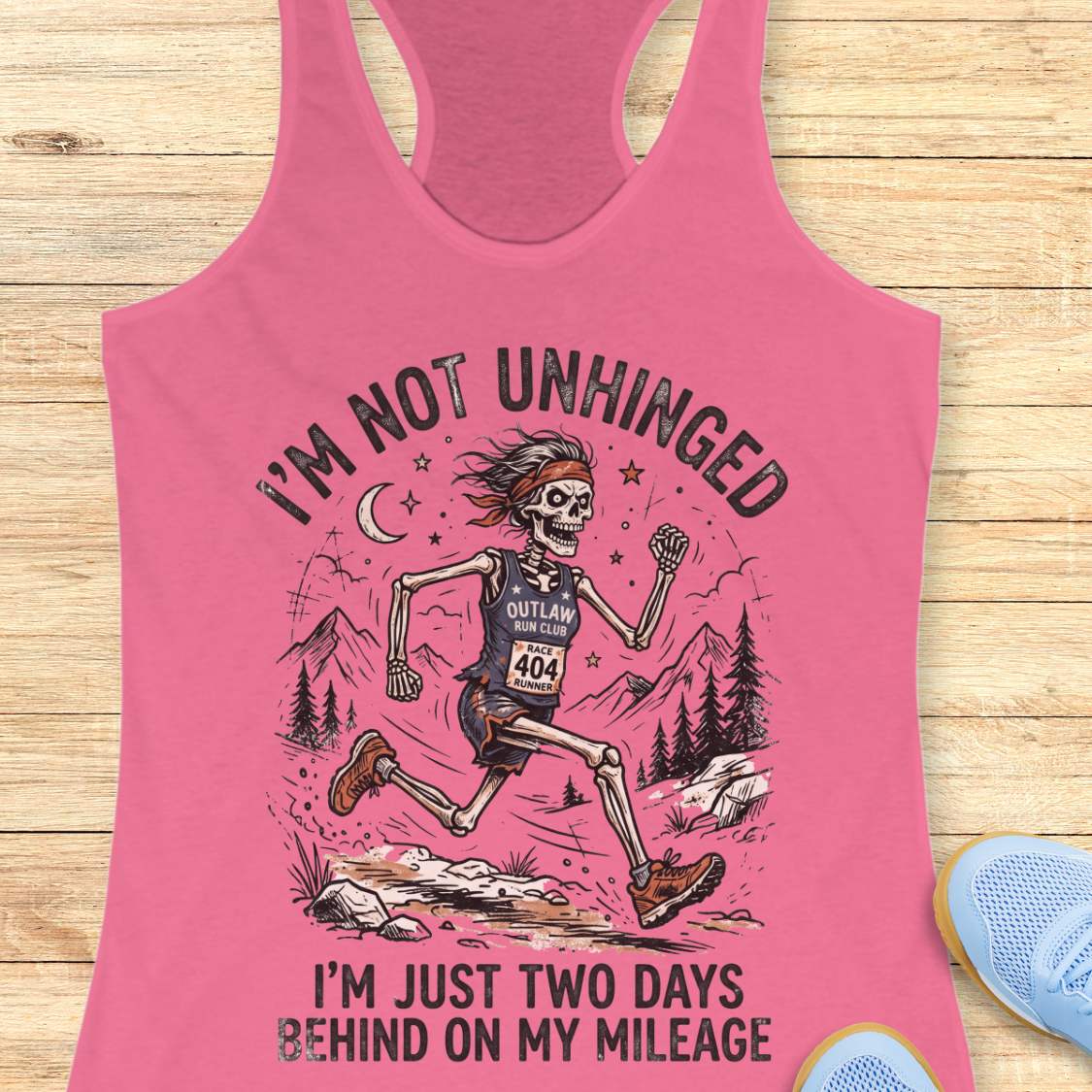 Pink tank top with skeleton graphic and text 'I'm not unhinged, I'm just two days behind on my mileage', on a wooden surface