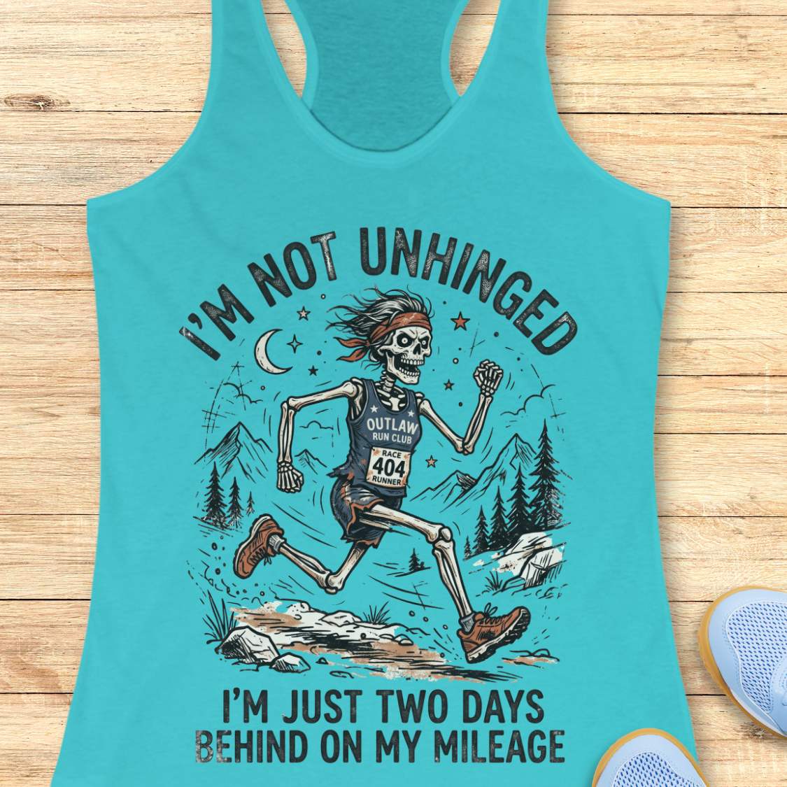 Turquoise tank top with skeleton graphic and text 'I'm not unhinged, I'm just two days behind on my mileage', on a wooden surface