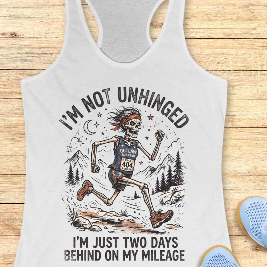 white tank top with skeleton runner graphic and text 'I'm not unhinged, I'm just two days behind on my mileage', on a wooden surface