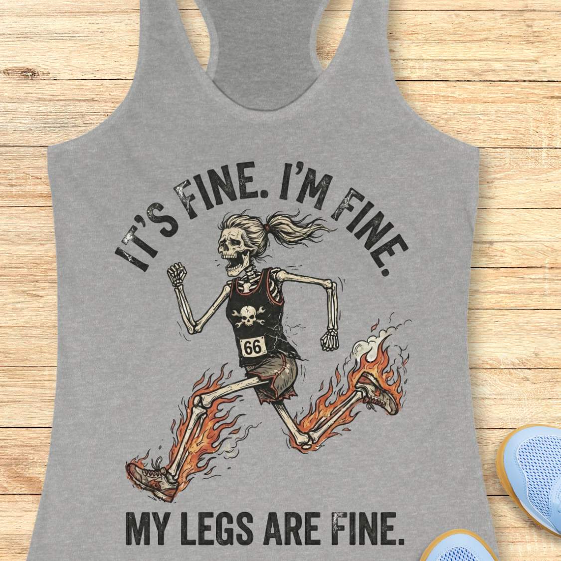 Gray tank top with skeleton design and text 'It's fine. I'm fine. My legs are fine.', on a wooden surface