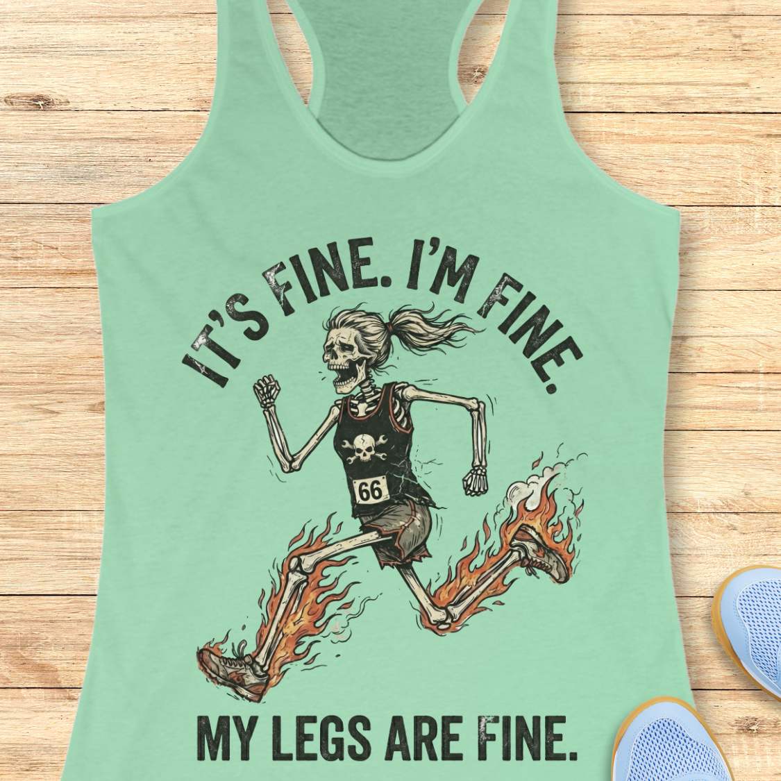 Mint green tank top with skeleton graphic and text  'It's fine. I'm fine. My legs are fine.',  on a wooden surface