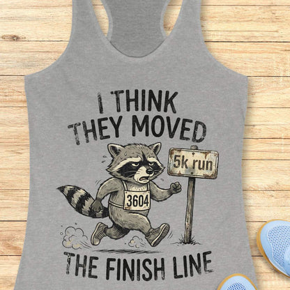 Gray tank top with a raccoon graphic and text 'I think they moved the finish line', on a wooden surface