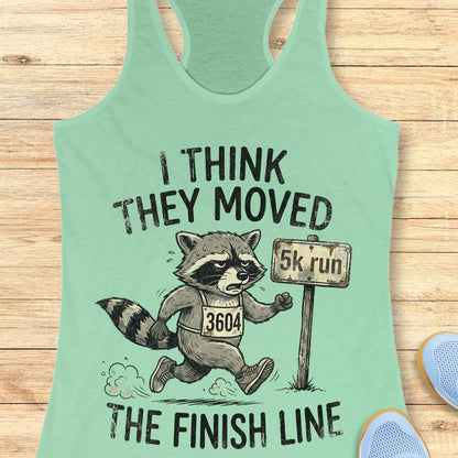 Mint green tank top with a raccoon graphic and text 'I think they moved the finish line', on a wooden surface