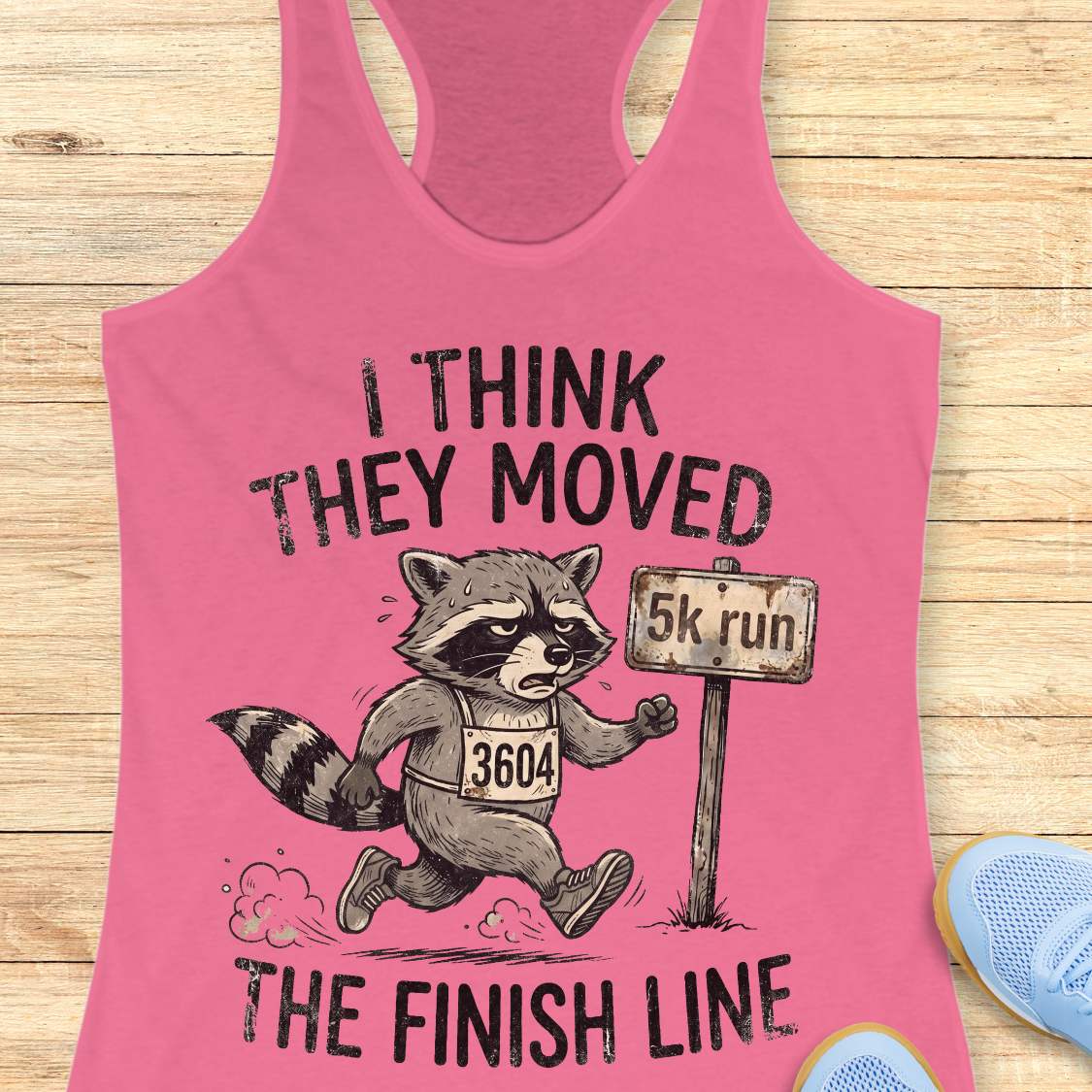 Pink tank top with a raccoon graphic and text 'I think they moved the finish line', on a wooden surface