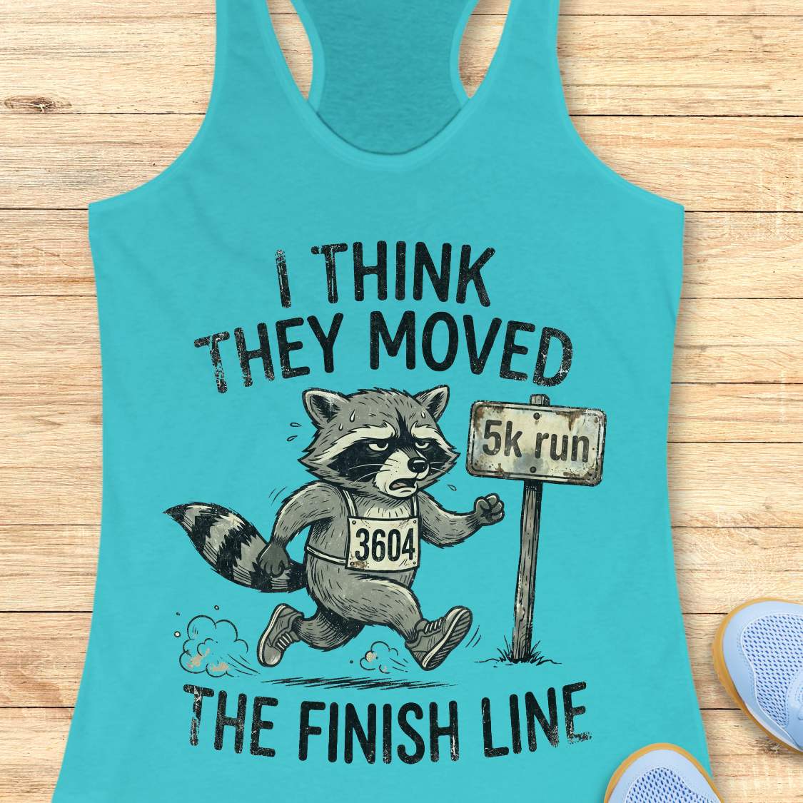 Turquoise tank top with raccoon graphic and text 'I think they moved the finish line', on a wooden surface