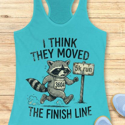 Turquoise tank top with raccoon graphic and text 'I think they moved the finish line', on a wooden surface