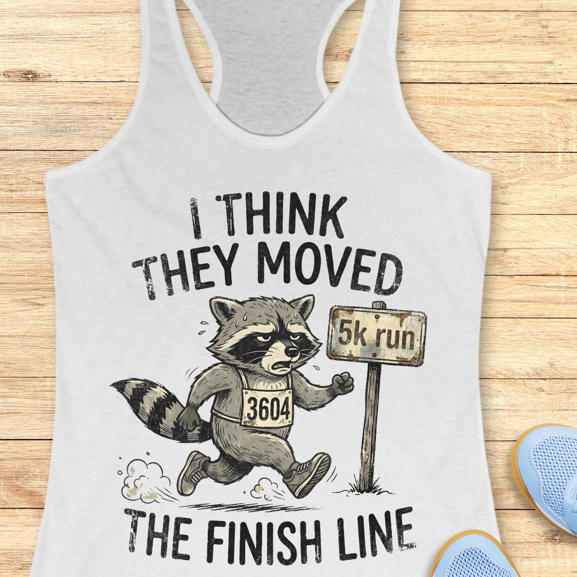 white tank top with a raccoon graphic and text 'I think they moved the finish line', on a wooden surface