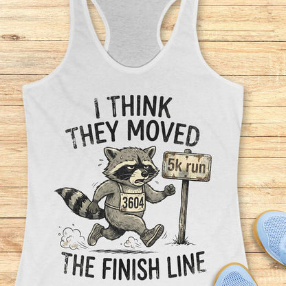 white tank top with a raccoon graphic and text 'I think they moved the finish line', on a wooden surface
