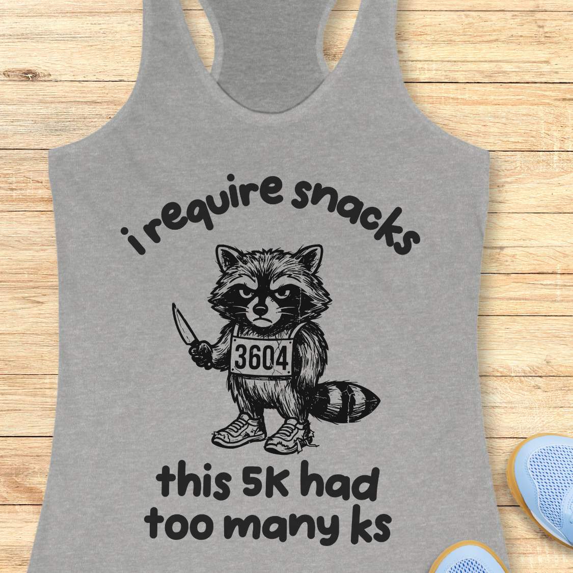 Gray tank top with a graphic of a raccoon and text 'i require snacks, this 5k had too many ks', on a wooden surface