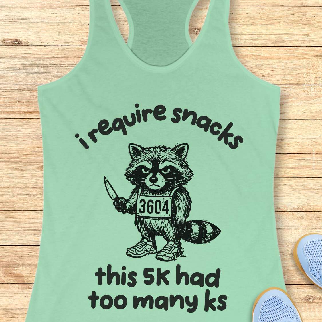 Mint green tank top with a graphic of a raccoon and text 'i require snacks, this 5k had too many ks', on a wooden surface