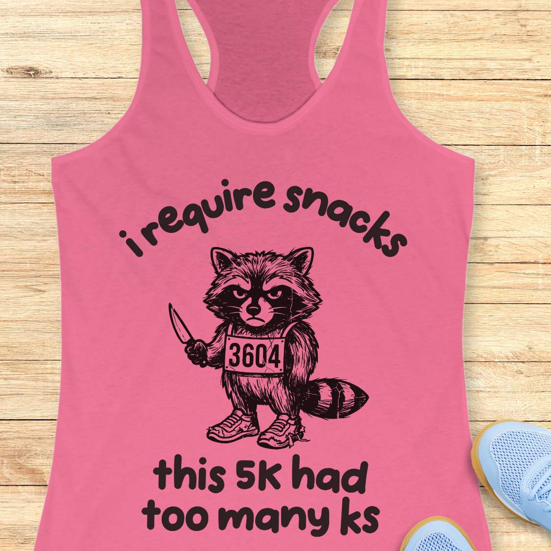 Pink tank top with a raccoon graphic and text 'i require snacks, this 5k had too many ks', on a wooden surface