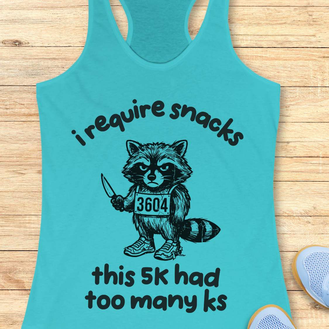 Turquoise tank top with a raccoon graphic and text 'i require snacks, this 5k had too many ks', on a wooden surface