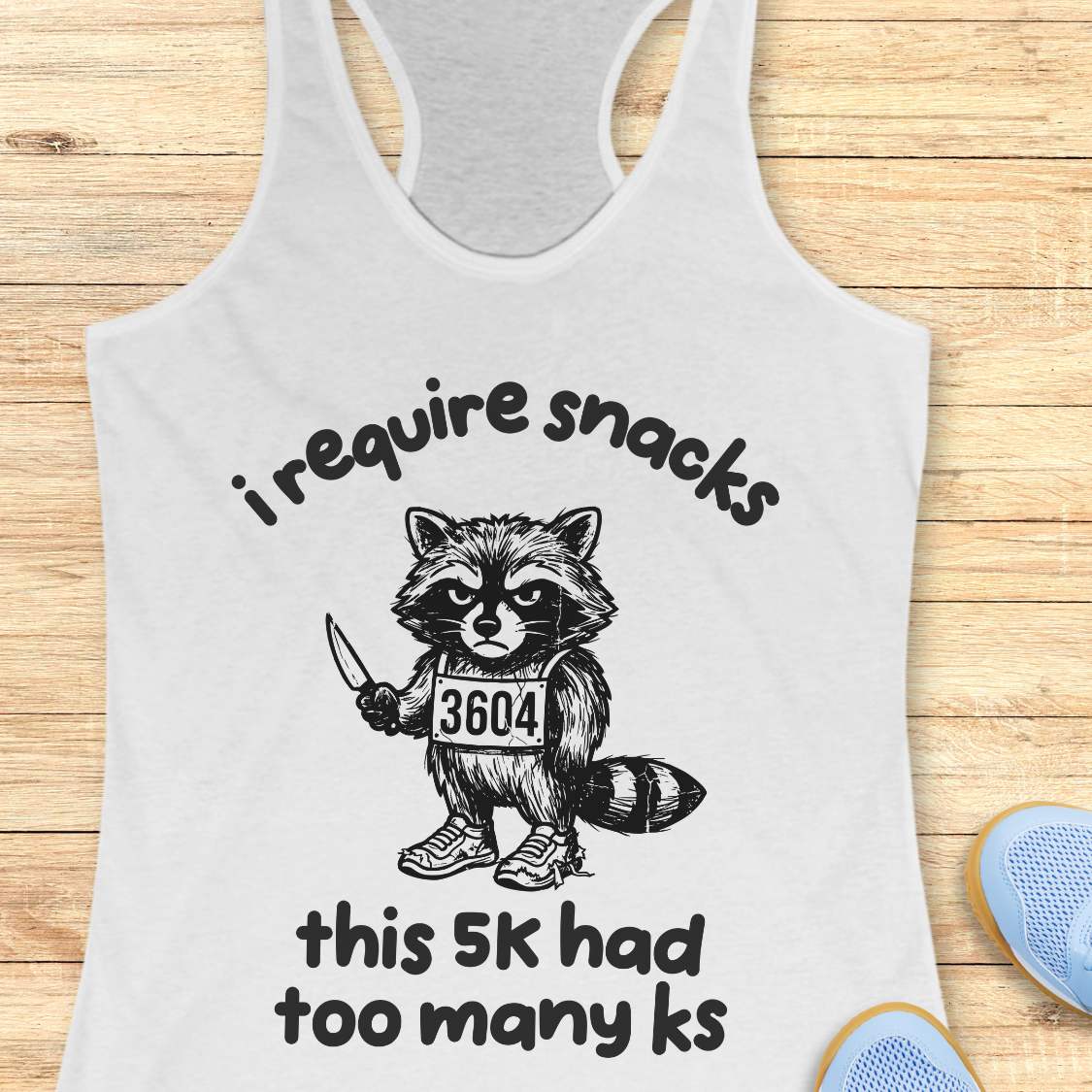 white tank top with a raccoon graphic and text 'i require snacks, this 5k had too many ks', on a wooden surface