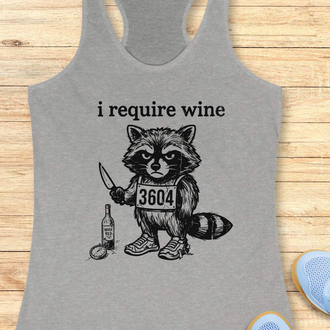 Gray tank top with a graphic of a raccoon holding a knife and bottle, text 'i require wine', on a wooden surface.