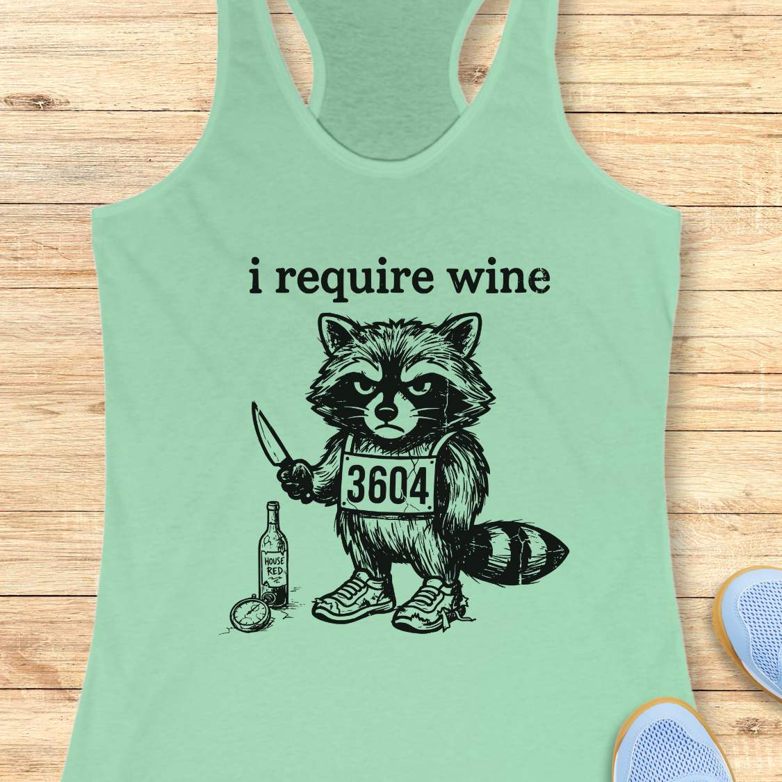 Light green tank top with a graphic of a raccoon holding a knife and a bottle, text 'i require wine', on a wooden surface.