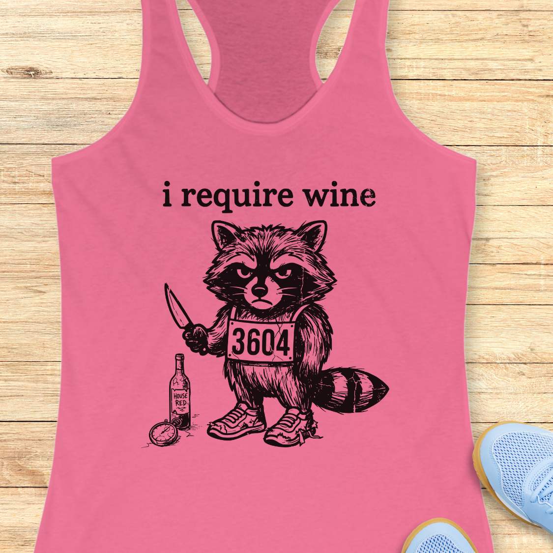 Pink tank top with a raccoon graphic and text 'i require wine' on a wooden surface.