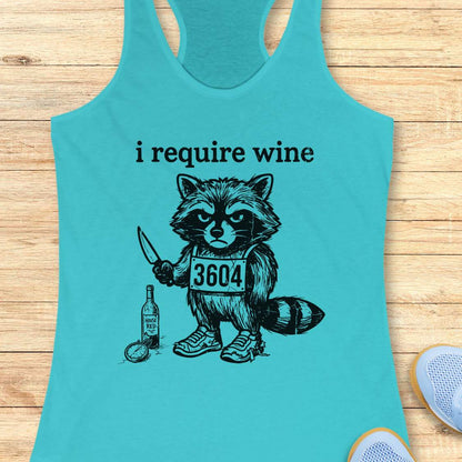 Turquoise tank top with a graphic of a raccoon holding a knife and a bottle, text 'i require wine', on a wooden surface.