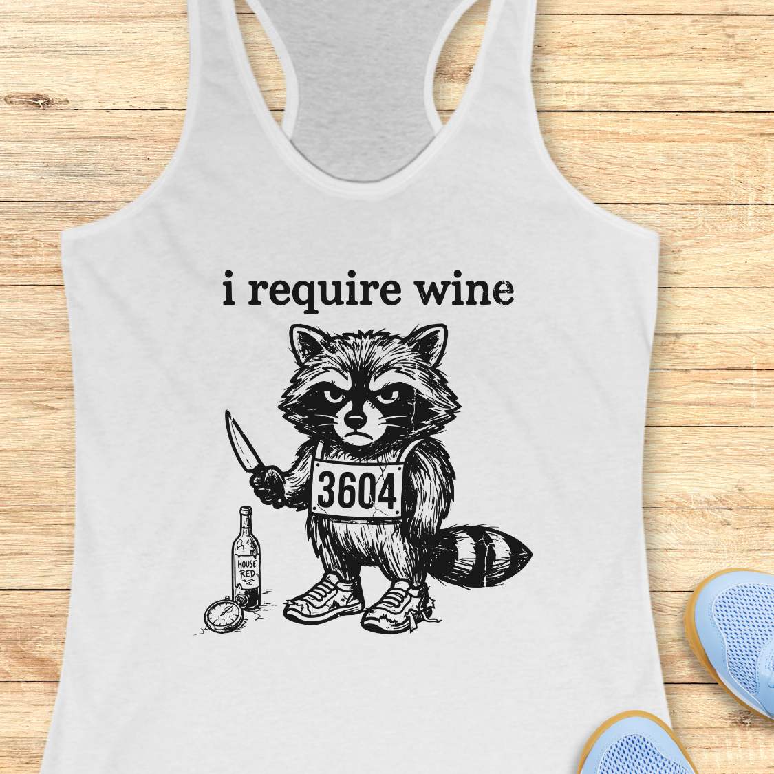 White tank top with a raccoon graphic and text 'i require wine' on a wooden surface.