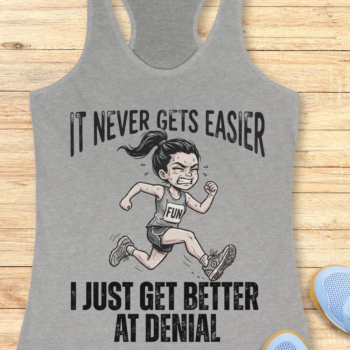 grey tank top with black graphic design and text 'it never gets easier, I just get better at denial', on a wooden surface