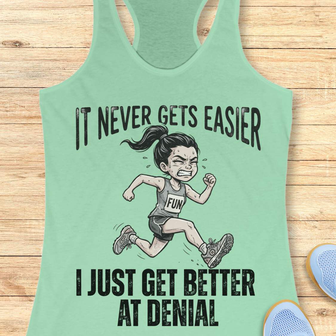 mint tank top with black graphic design and text 'it never gets easier, I just get better at denial', on a wooden surface