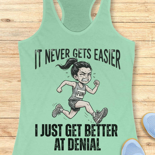 mint tank top with black graphic design and text 'it never gets easier, I just get better at denial', on a wooden surface