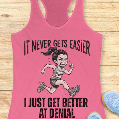 pink tank top with black graphic design and text 'it never gets easier, I just get better at denial', on a wooden surface