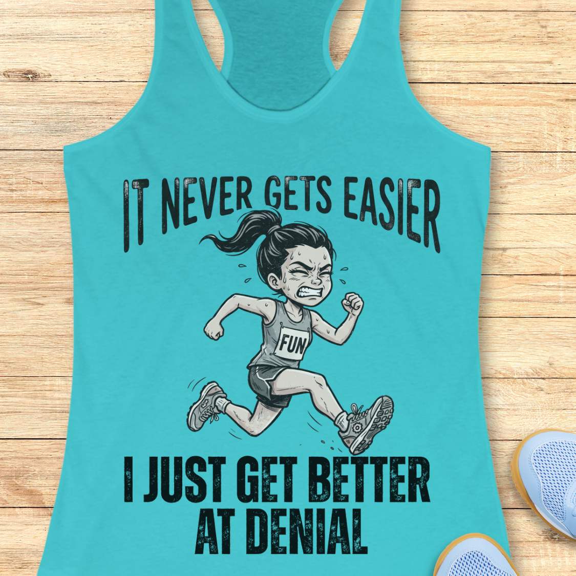 blue tank top with black graphic design and text 'it never gets easier, I just get better at denial', on a wooden surface